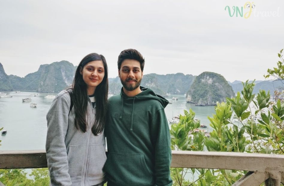 A young couple posing at a scenic viewpoint overlooking Ha Long Bay, with limestone islands and cruise boats in the background.