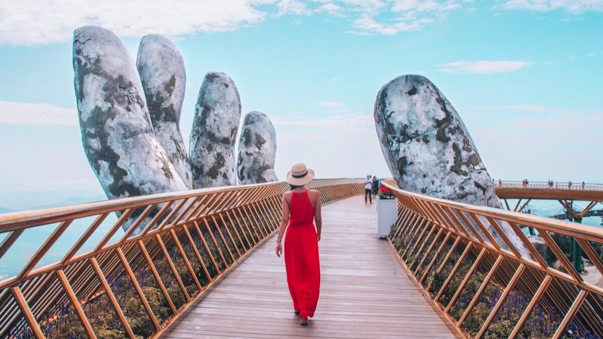 A woman in a red dress walks across the iconic Golden Bridge at Ba Na Hills, Da Nang, Vietnam, with the famous giant stone hands and panoramic mountain views.