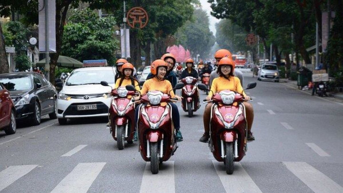A group of foreign tourists wearing helmets and riding scooters along a tree-lined street, taking part in a guided motorbike tour in Hanoi.
