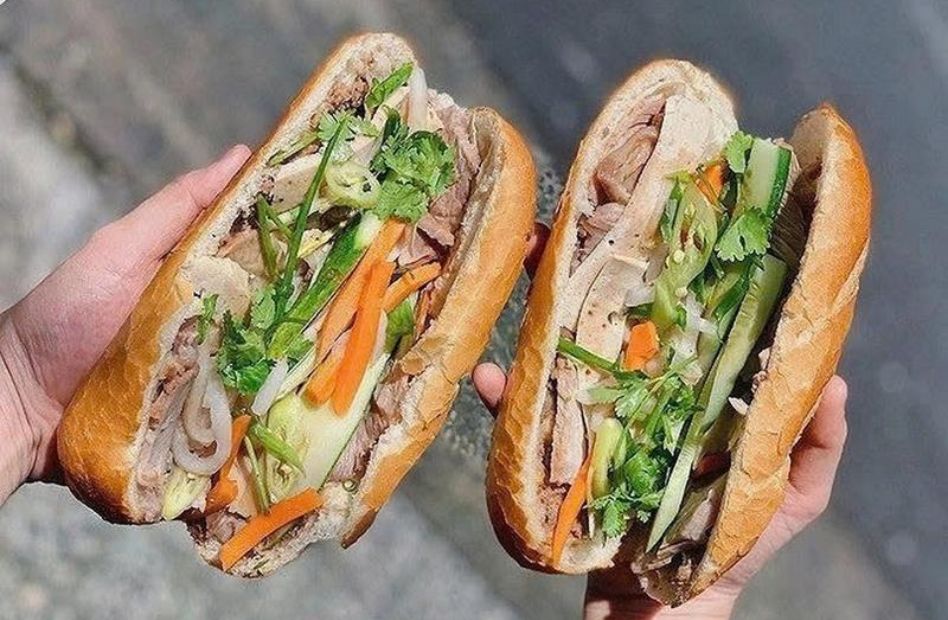 Two freshly made Hoi An banh mi sandwiches filled with sliced pork, pickled vegetables, cucumber, cilantro, and savory sauces in crispy golden baguettes.
