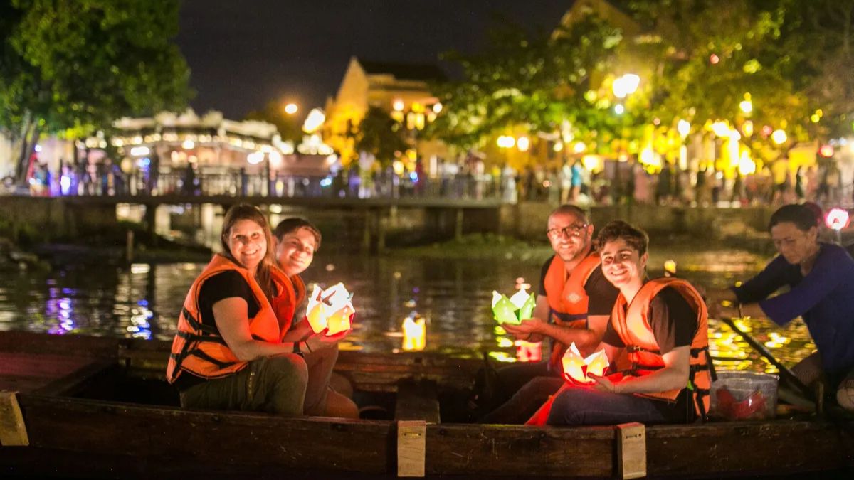 International tourists wearing life jackets release colorful lanterns while enjoying a traditional boat ride on the Hoai River in Hoi An at night.