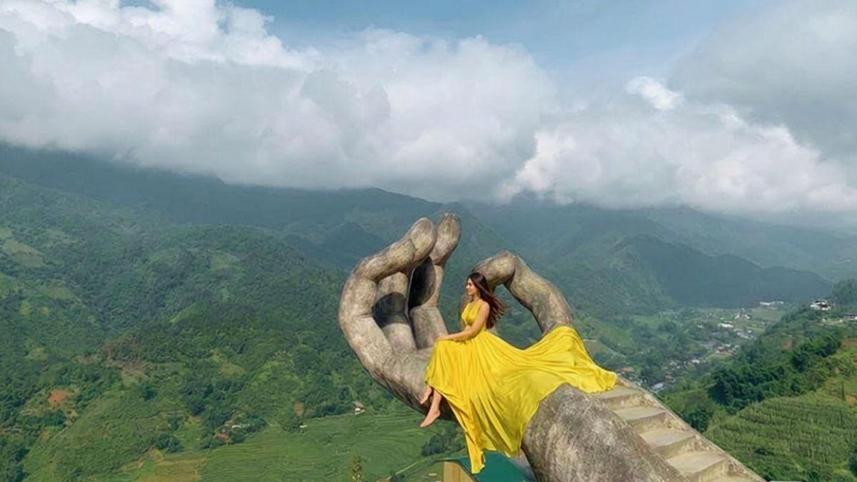 A woman in a flowing yellow dress poses on the giant hand sculpture at Moana Sapa, overlooking the stunning mountains of Sapa, northern Vietnam.