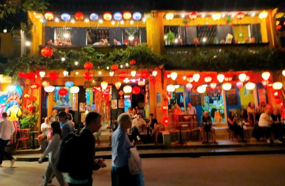 People walking along a street in Hoi An Ancient Town at night in front of traditional yellow heritage buildings decorated with glowing colorful silk lanterns and outdoor seating for cafes.