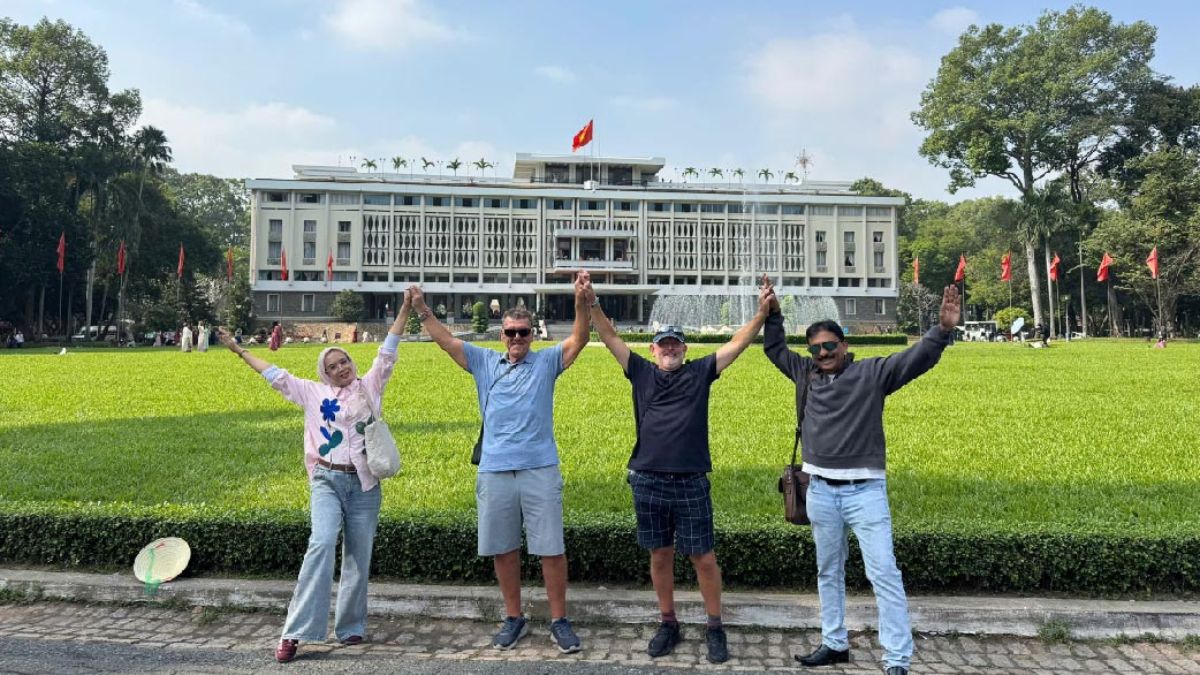 Group of tourists posing with raised hands in front of the Independence Palace (Reunification Palace) in Ho Chi Minh City, Vietnam.