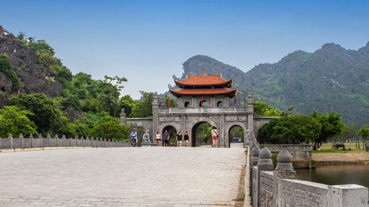 A wide stone bridge leading to a traditional three-arched stone gate with a double-tiered red-tiled roof, set against lush green limestone mountains and a clear blue sky at the Hoa Lu Ancient Capital (Dinh and Le Dynasties temples) in Ninh Binh, Vietnam.