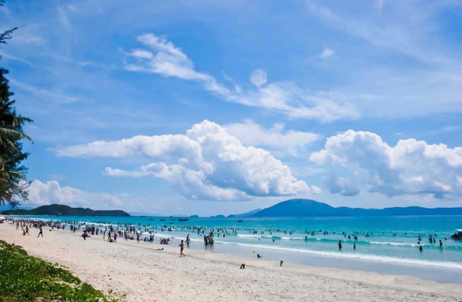 Golden sandy shoreline at Do Son Beach in Hai Phong, where gentle waves meet the coast as visitors relax, swim, and enjoy the scenic seaside surrounded by lush green hills.