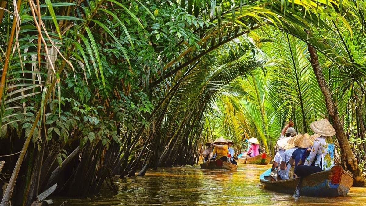 Tourists riding traditional sampan boats through a peaceful coconut canal in the Mekong Delta, surrounded by lush nipa palm forests in southern Vietnam.