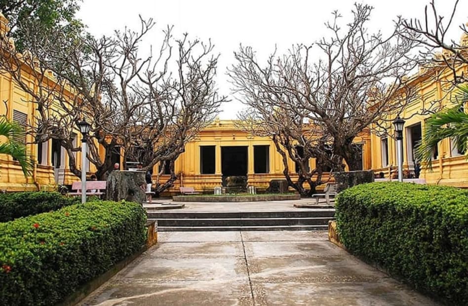 A wide courtyard view of the yellow French-colonial building of the Museum of Cham Sculpture in Da Nang, Vietnam, framed by green trimmed hedges, paved walkways, and large frangipani trees with bare branches.