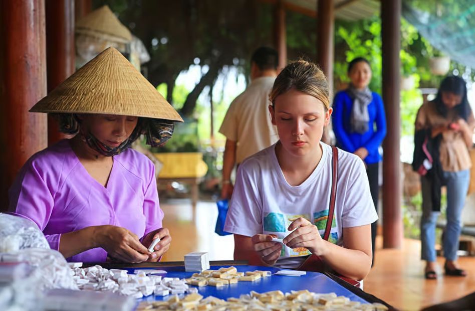 A local Vietnamese woman wearing a traditional conical hat teaches a young woman how to make a coconut-based craft at a workshop in Ben Tre.